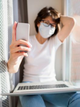 Pretty woman in medical mask remote works from home. She is making selfie on window sill with laptop on knees. Lockdown quarantine because of coronavirus COVID19. Self isolation at home.