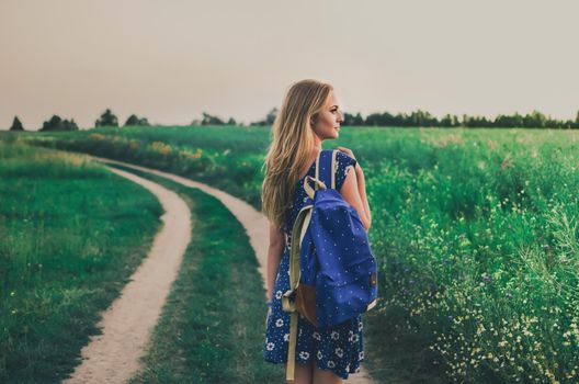 young thin blonde woman stands in dress and backpack on her shoulder in middle of green field On walking path in summer at sunset