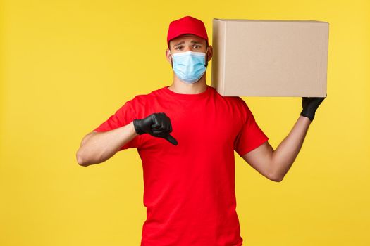 Express delivery during pandemic, covid-19, safe shipping, online shopping concept. Upset and disappointed courier in red uniform, medical mask, show thumb-down displeased, holding package.