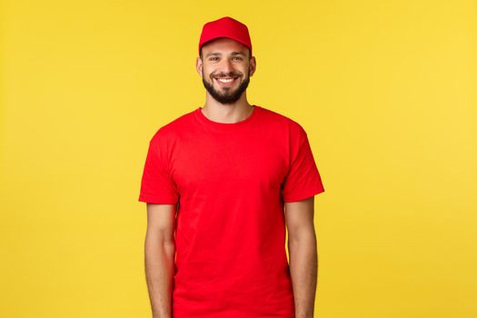 Express mail delivery, package transfer and logistics concept. Handsome friendly-looking courier in red uniform of company, t-shirt and cap, smiling broadly as standing yellow background.