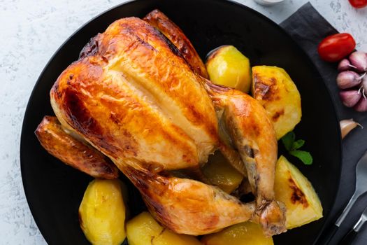 Grilled whole chicken in a black plate on white table, baked meat with potatoes. Top view, close up