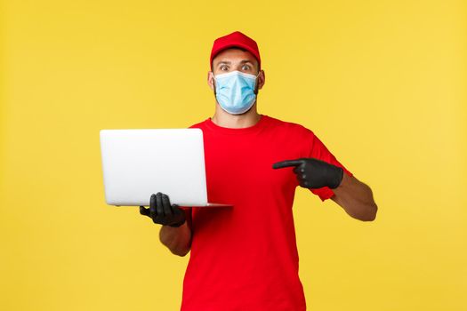 covid-19, safe shopping, express delivery and order tracking concept. Surprised courier or employee in red uniform, medical mask, pointing finger at laptop with impressed excited face.