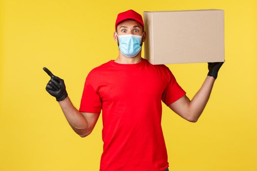 Express delivery during pandemic, covid-19, safe shipping, online shopping concept. Enthusiastic courier in red uniform, medical mask and gloves, hold box on shoulder, pointing finger left.
