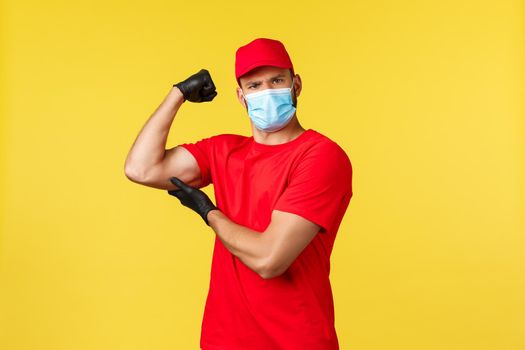 Express delivery during pandemic, covid-19, safe shipping, online shopping concept. Strong and determined courier in red uniform, medical mask and gloves, flex biceps, brag with muscles.