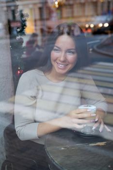 A beautiful girl sits in cafe and looks out window thoughtfully. Reflection of city in window. Smiling brunette woman with long hair drinks cappuccino coffee, vertical