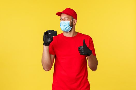 Express delivery in pandemic, covid-19, safe online shopping concept. Friendly smiling courier in red uniform, medical mask and gloves, advice use credit cards during coronavirus instead cash.