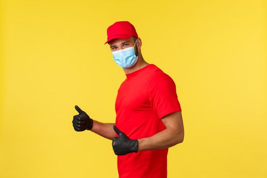 Express delivery during pandemic, covid-19, safe shipping, online shopping concept. Handsome smiling courier in red uniform, medical mask and PPE gloves, thumb-up guarantee safe and fast shipment.