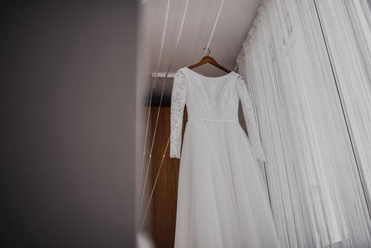 A white luxurious beautiful wedding dress hangs on a hanger. Pleated white chiffon curtains hang around.