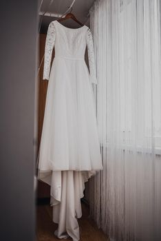 A white luxurious beautiful wedding dress hangs on a hanger. Pleated white chiffon curtains hang around.