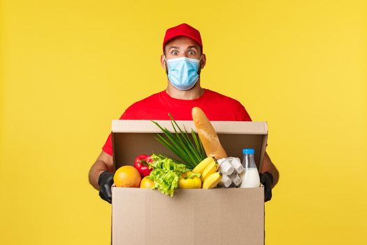 Takeaway delivery, covid-19 quarantine and groceries concept. Surprised courier in medical mask, uniform red cap and t-shirt, staring shocked, bring food order box to customer home, yellow background.