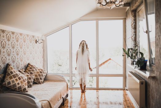 a lady with a veil on her head in white lace underwear goes to the window taking off her silk robe Throwing it on the ground. Orange brown parquet floor. Morning of the bride.