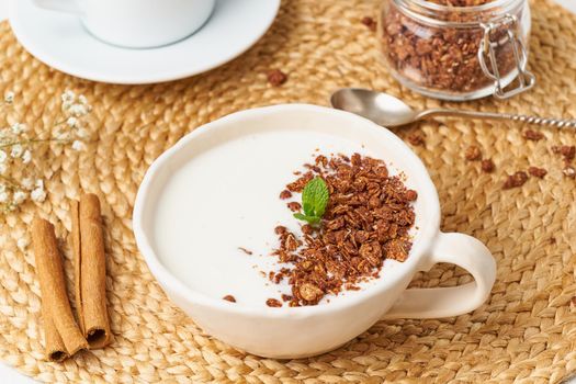 Yogurt with chocolate granola in cup, breakfast with tea on beige background, side view, close up