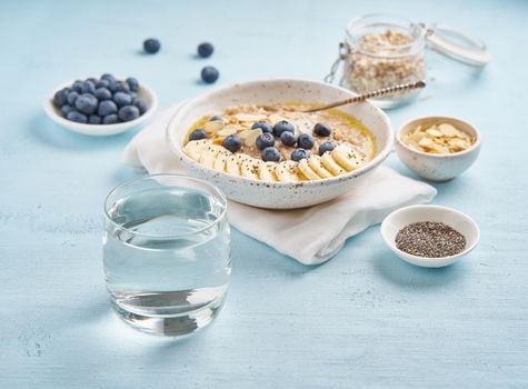 Glass of clean water and healthy diet breakfast with oatmeal, blueberries, banana on blue light background. Side view.