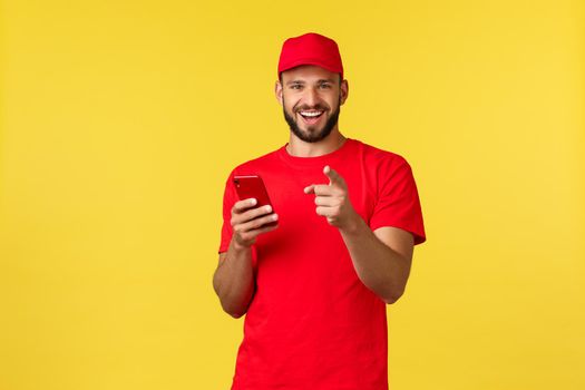 Contactless payment, online delivery, internet shopping and parcel tracking concept. Handsome bearded employee, courier guy in red t-shirt and cap, pointing at camera gotcha as using mobile phone.