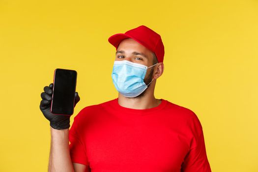 Covid-19, delivery orders, shopping, contactless payment and social distancing concept. Handsome courier in red uniform and medical mask, gloves, showing smartphone app, mobile phone screen.