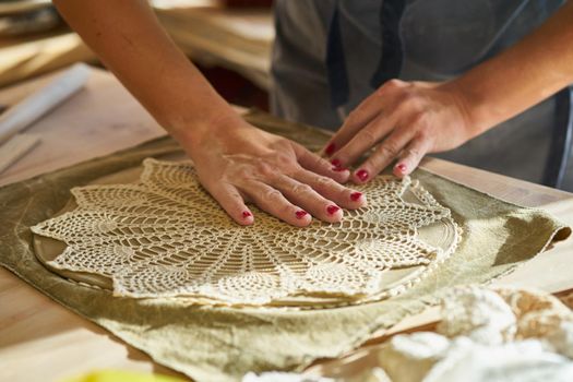 Woman making pattern on ceramic plate, hands close-up, focus on palms with clay and lace. Creative hobby concept