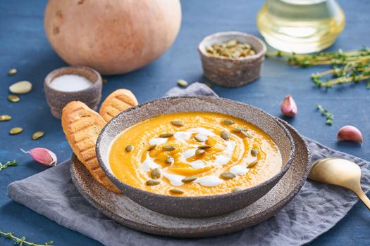 Pupmkin cream soup puree on dark bright blue wooden table, seeds, toast, dietary vegetarian lunch, side view