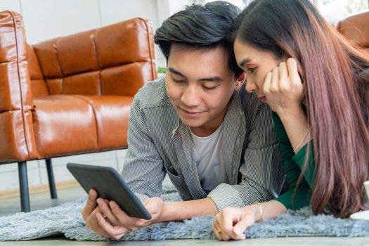 Happy Asian couple use tablet while lying down on carpet at living room floor. Love and relationship concept.