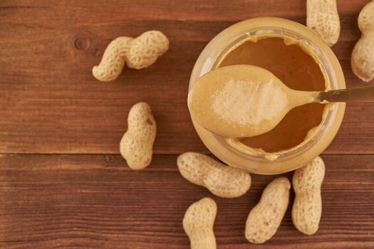 jar of peanut butter and peanuts in shell on brown wooden table, side view, fresh crushed ground nuts, top view, copy space