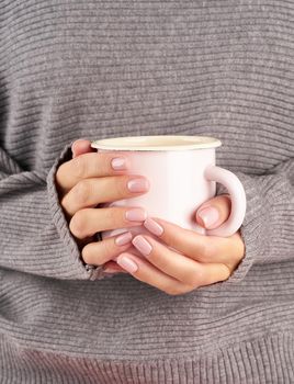 morning hot coffee at work in office on a cold autumn morning, hands holding a mug with a drink, gray sweater, pink manicure, close up, vertical