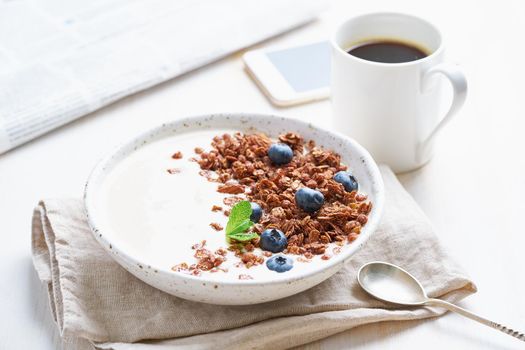 Yogurt with chocolate granola, bilberry. Breakfast with cup of coffee, mobile, newspaper on a white background, side view