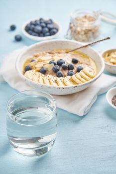 Glass of clear water and healthy diet breakfast with oatmeal, blueberries, banana on blue light background. Vertical. Side view.