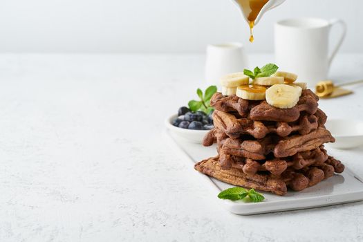 Chocolate banana waffles with maple syrup on white table, copy space, side view. Sweet brunch, a maple syrup drop.