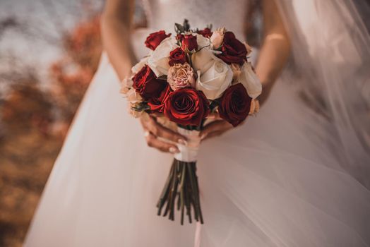 A bride in a white wedding dress holds a bouquet of red and white roses in front of her. Autumn orange foliage on the background.