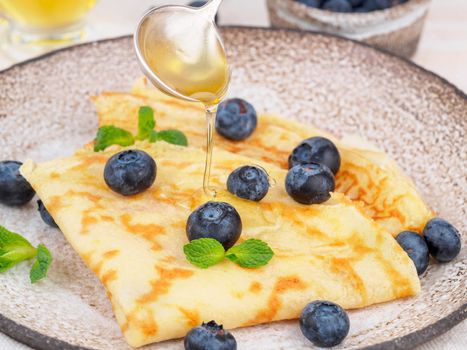 Two pancakes with blueberries and honey on plate, honey flows from spoon, side view, close-up