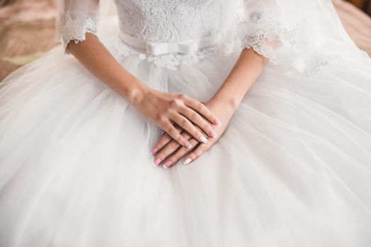 The female hands of the fair-skinned bride lie on the white wedding dress. A ring on the hand of a young girl.