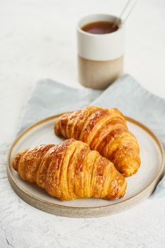 Two delicious croissants on a plate and a hot drink in a mug. Morning French breakfast with fresh pastries. Light gray background, vertical