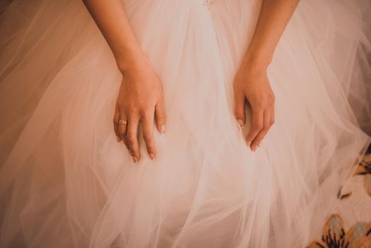 The female hands of the fair-skinned bride lie on the white wedding dress. A ring on the hand of a young girl.