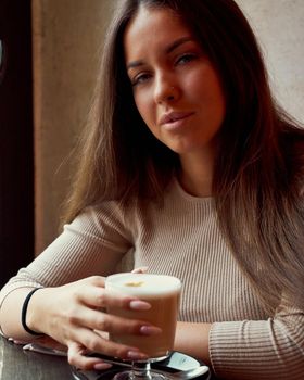 Beautiful pensive happy girl sitting in cafe in Christmas holidays, smiling and dreaming. Brunette woman with long hair drinks cappuccino coffee, latte and looks to camera, vertical