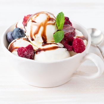 Balls of vanilla ice cream in mug with raspberry and blueberries, chocolate syrup topping. Side view summer dessert, white, bright background, macro