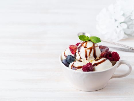 Balls of vanilla ice cream in mug with raspberry and blueberries, chocolate syrup topping. Side view, copy space, white bright background
