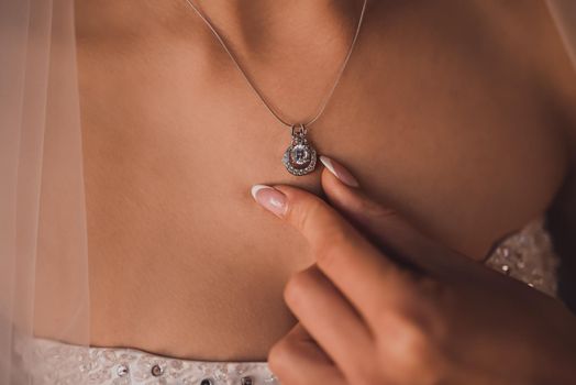 Silver gemstone pendant around the neck of a tanned bride. The girl holds the pendant by fingers with manicure.