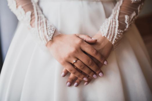 The female hands of the fair-skinned bride lie on the white wedding dress. A ring on the hand of a young girl.