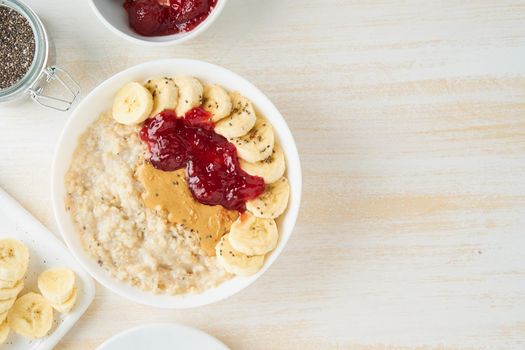Oatmeal porridge with strawberry jam, peanut butter, banana. Top view, copy space on white wooden light background, healthy vegan diet breakfast