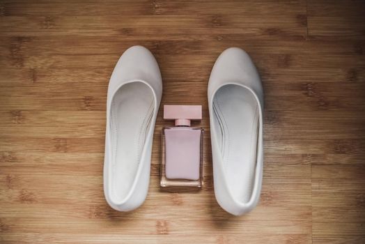 between two white shoes are pink women's perfumes, eau de toilette. light beige women's high-heeled shoes on the floor on a wooden brown background. classic shoes of the bride.