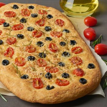 Focaccia, pizza, italian flat bread with tomatoes, olives and a rosemary on brown table, side view
