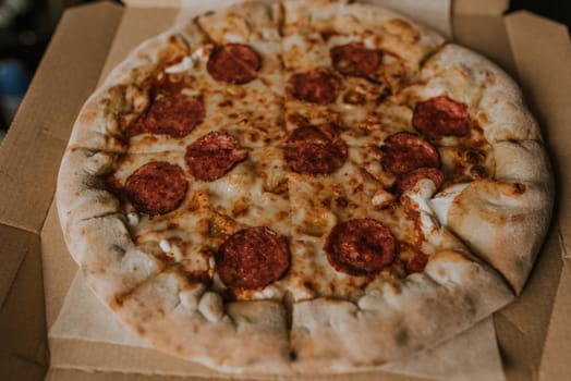 A large round juicy mouth-watering pizza with cheese and salami lies on a cardboard box from a delivery package. paper backing. close view. see flour. good thin dough