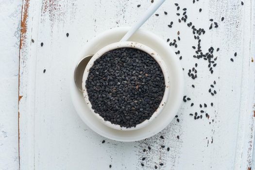 Black Sesame on a white bowl with spoon. White wooden background. Top view, close up. Copy space.