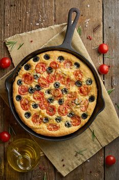 Focaccia, pizza in skillet, italian flat bread with tomatoes, olives and a rosemary. Vertical, wooden table