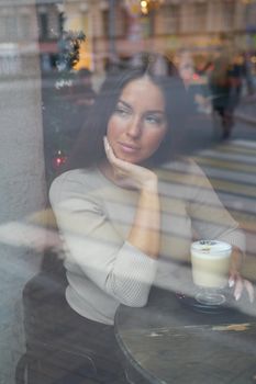 A beautiful girl sits in cafe and looks out window thoughtfully. Reflection of city in window. Brunette woman with long hair drinks cappuccino coffee, vertical