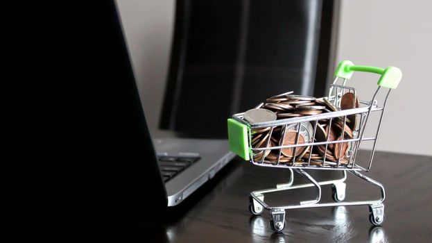 Shopping cart full of money and laptop on a wooden desktop: e-commerce, e-payments and online shopping concept