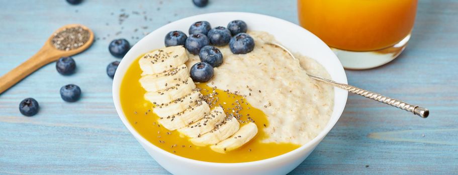 Banner with oatmeal, bananas, blueberries, chia seeds, mango jam on blue wooden background. Side view. Healthy breakfast
