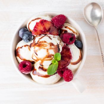 Balls of vanilla ice cream in mug with raspberry and blueberries, chocolate syrup topping. Top view summer dessert, white, bright background, macro