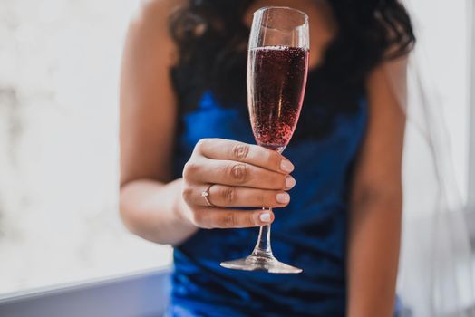 A black-haired, curly-haired, tanned bride dressed in blue satin suede pajamas with a ring on her ring finger holds a transparent crystal glass filled with red champagne.