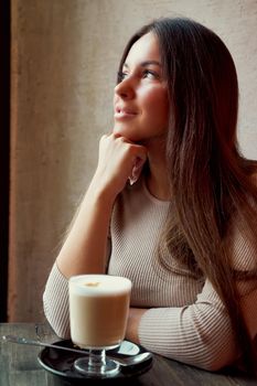 Beautiful pensive happy girl sitting in cafe in Christmas holidays, smiling and dreaming. Brunette woman with long hair drinks cappuccino coffee, latte and looks out window, vertical, dark backdrop