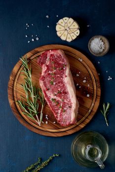 Big whole piece of raw beef meat, striploin, olive oil on a wooden cutting board on dark blue background. Seasoning steak with salt, vertical